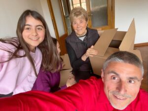 Moving to a new home concept. Young girl and her parents and grandmother seated near cardboard boxes along the parquet floor taking selfies with smartphone.